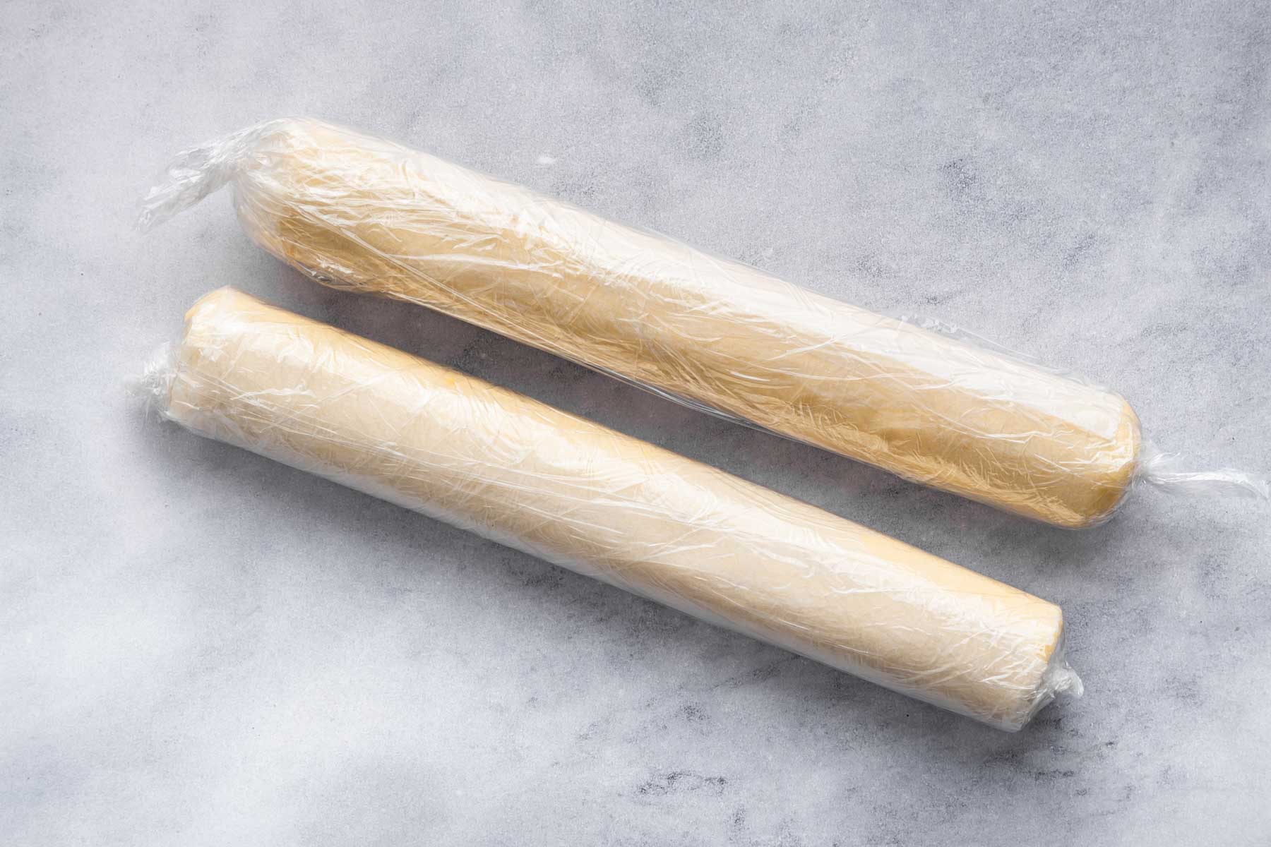Two skinny logs of dough rolled and wrapped on a marble counter.