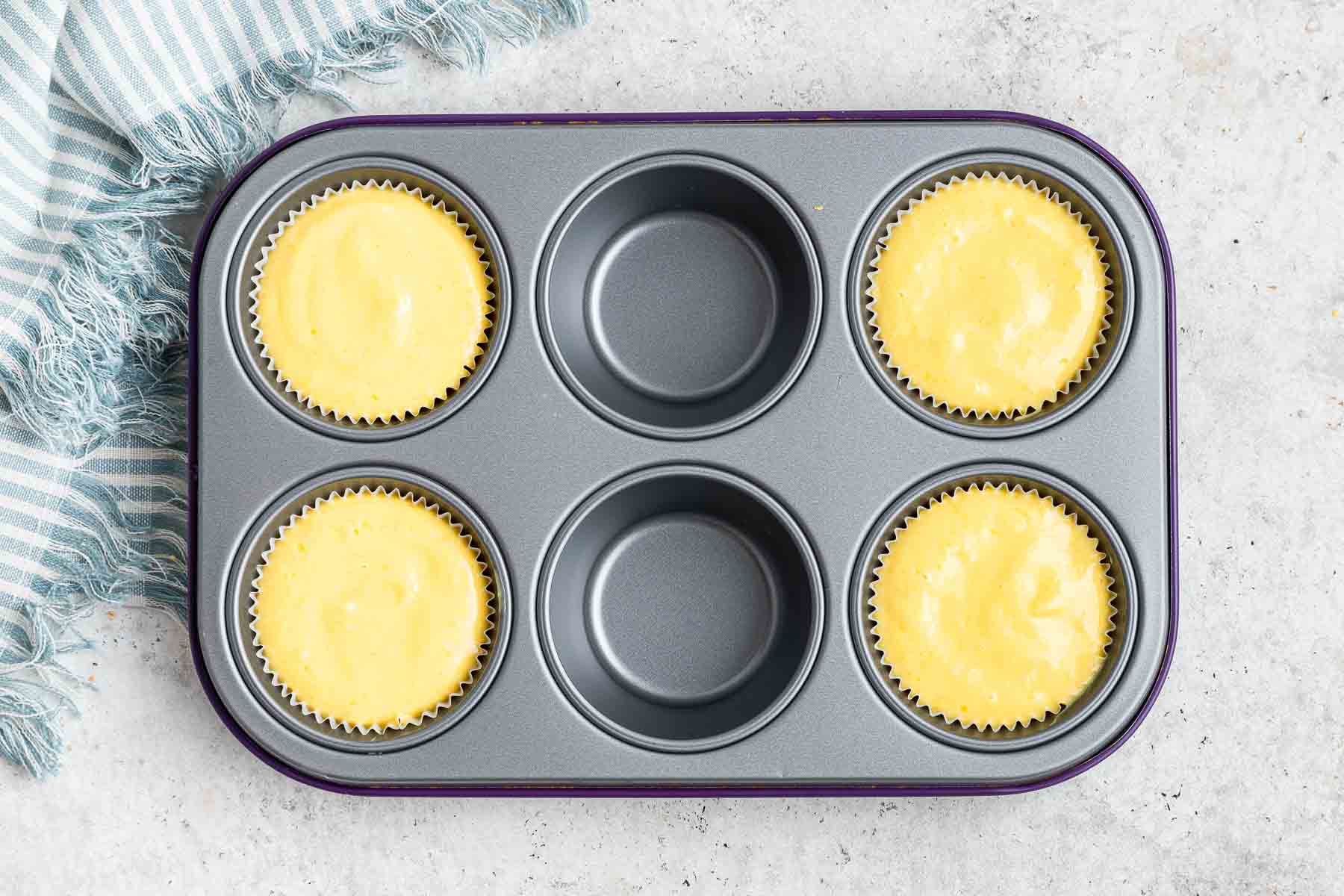 Muffin pan with four cups full of yellow batter with blue kitchen towel on side.