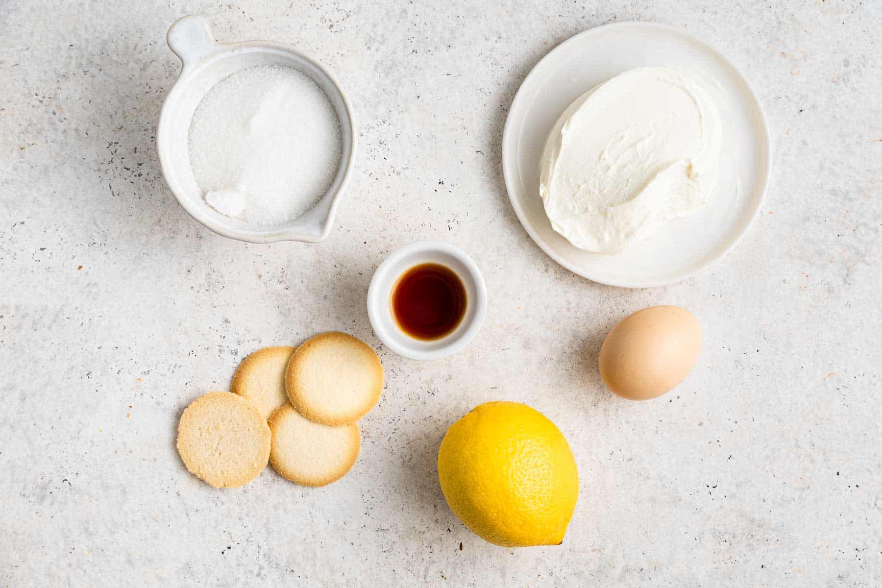 Bowl of sugar, cream cheese, lemon, egg and vanilla extract on grey counter.