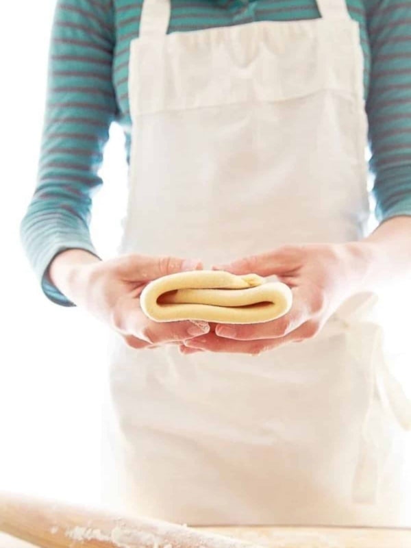Woman in blue shirt and white apron holding folded puff pastry recipe.