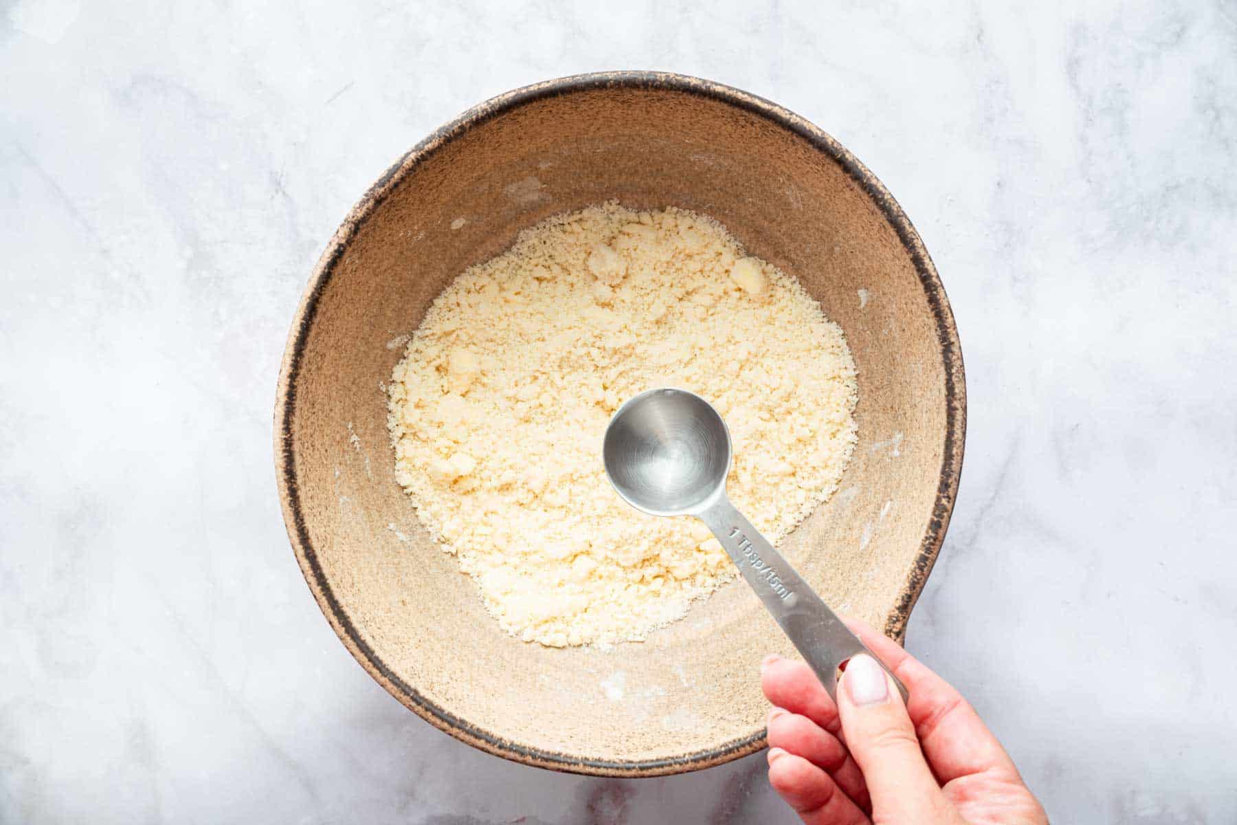Hand adding one teaspoon of clear liquid to bowl of crumbly pastry.