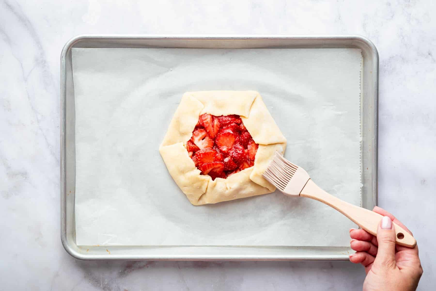 Hand brushing milk on pastry crust with berries in center.