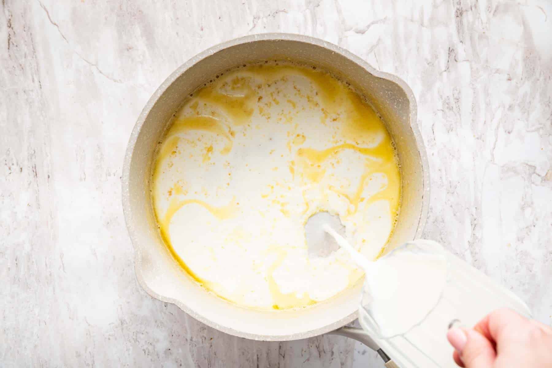 Hand pouring milk into roux mixture in saucepan.