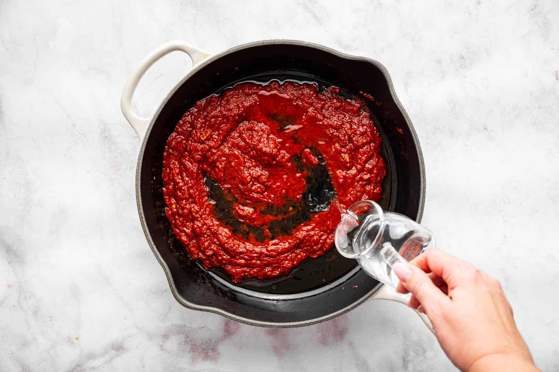 Hand pouring clear liquid into black skillet with tomato sauce.